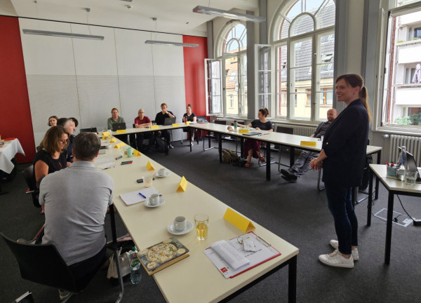 Trainerin lacht den Fortbildungsteilnehmern, die in Hufeisenform sitzen, zu